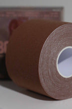 Close-up image of a roll of Fixie Boob Tape in the dark brown Hickory color. The focus is on the finely woven, textured surface of the dark brown-tone fabric tape, tightly wound in concentric layers around a white cardboard core. The background is softly blurred, with hints of product packaging visible.