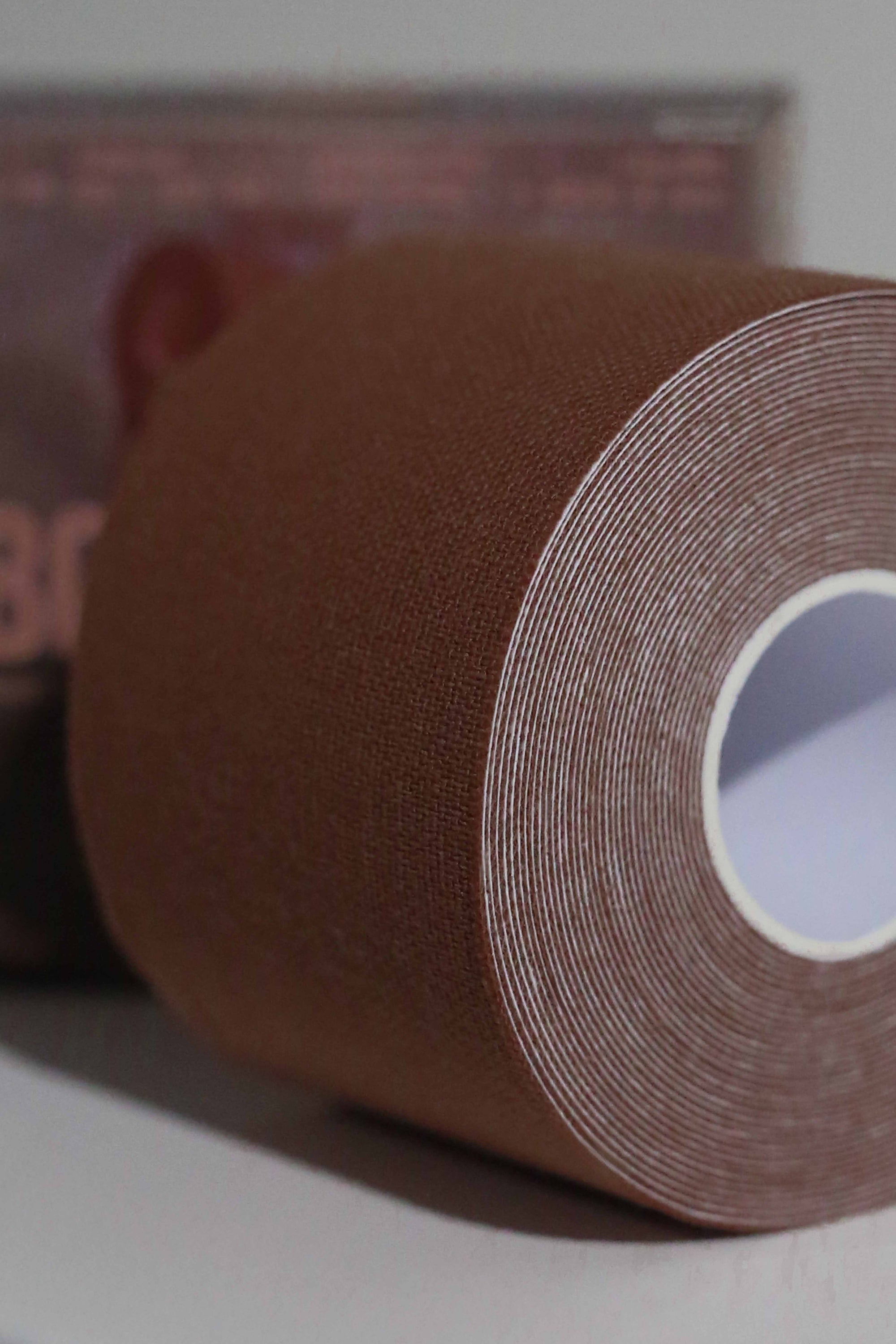 Close-up image of a roll of Fixie Boob Tape in the dark brown Hickory color. The focus is on the finely woven, textured surface of the dark brown-tone fabric tape, tightly wound in concentric layers around a white cardboard core. The background is softly blurred, with hints of product packaging visible.