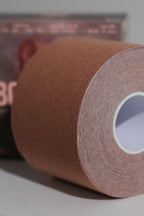 Close-up image of a roll of Fixie Boob Tape in the Sienna color. The focus is on the finely woven, textured surface of the brown-tone fabric tape, tightly wound in concentric layers around a white cardboard core. The background is softly blurred, with hints of product packaging visible.