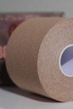 Close-up image of a roll of Fixie Boob Tape in the Winter Blossom color. The focus is on the finely woven, textured surface of the nude-tone fabric tape, tightly wound in concentric layers around a white cardboard core. The background is softly blurred, with hints of product packaging visible.